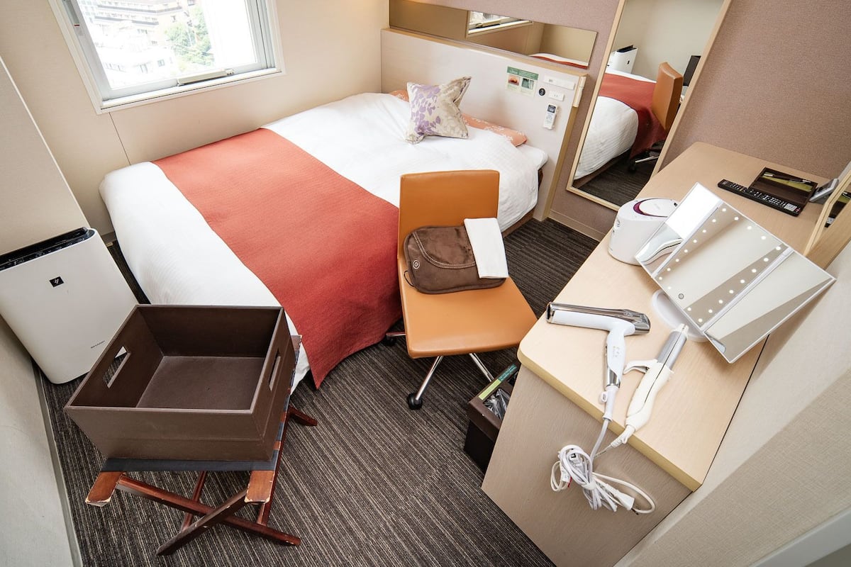 Una habitación de hotel con una cama, un escritorio con espejo, un cesto de basura pequeño y una maleta.