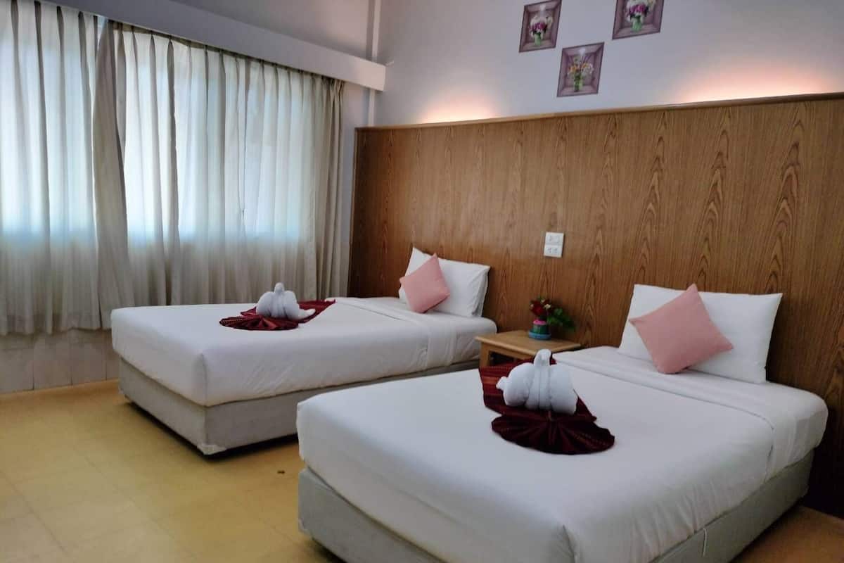 A hotel room with two beds, each with a white bedspread and a red blanket. There is a wooden headboard, a small table with a vase of flowers, and a window with white curtains.