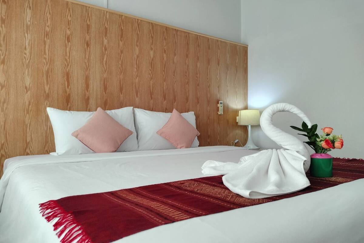 A hotel room with a wooden headboard, a bed with white and pink pillows, a red throw blanket, a white towel, and a vase with flowers.