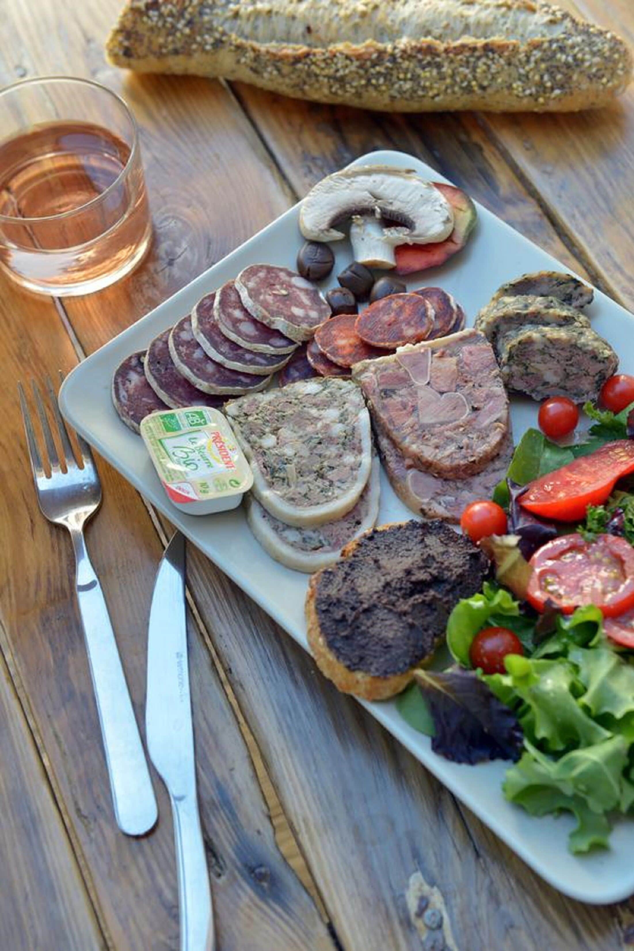 Un plato con diversas carnes, una bebida en un vaso y una baguette sobre una mesa de madera.