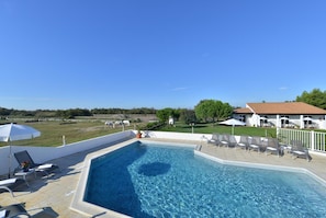 Ein rechteckiger Swimmingpool mit klarem, blauem Wasser, umgeben von Liegestühlen und Sonnenschirmen, mit einem weißen Gebäude und einer Rasenfläche im Hintergrund.