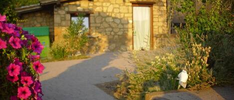 A stone building with a paved entrance, surrounded by greenery and flowering plants.