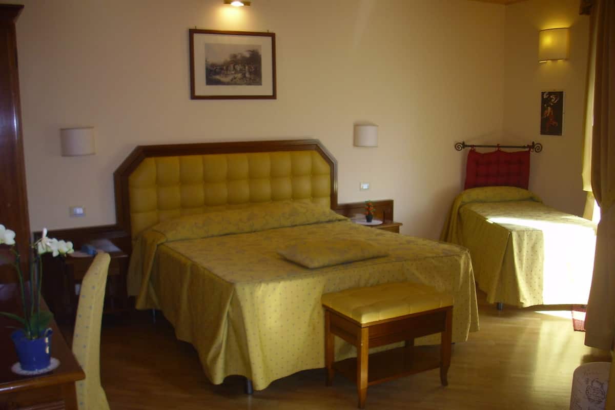 A hotel room with a large bed, a smaller bed, a wooden bench, a table with a vase of flowers, and a framed picture on the wall.