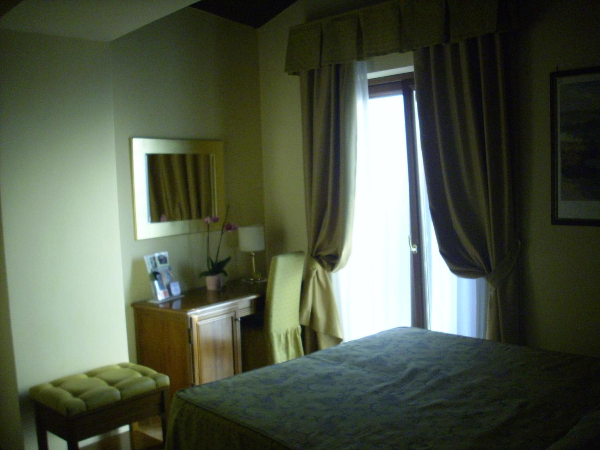 A hotel room with a bed, a desk with a lamp, a chair, a small table, and a potted plant.