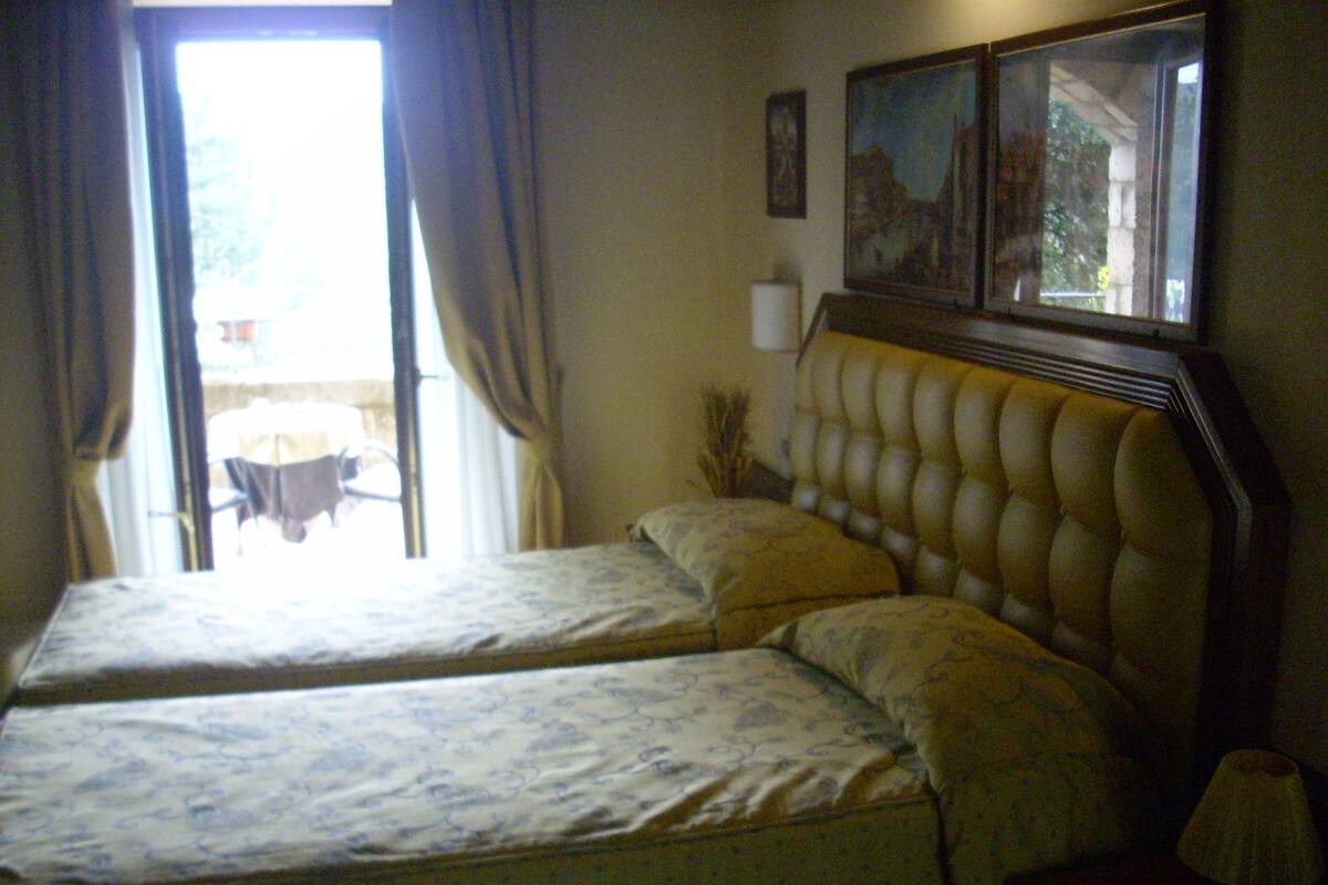 A room with two beds, a window with curtains, a lamp, and a painting on the wall.