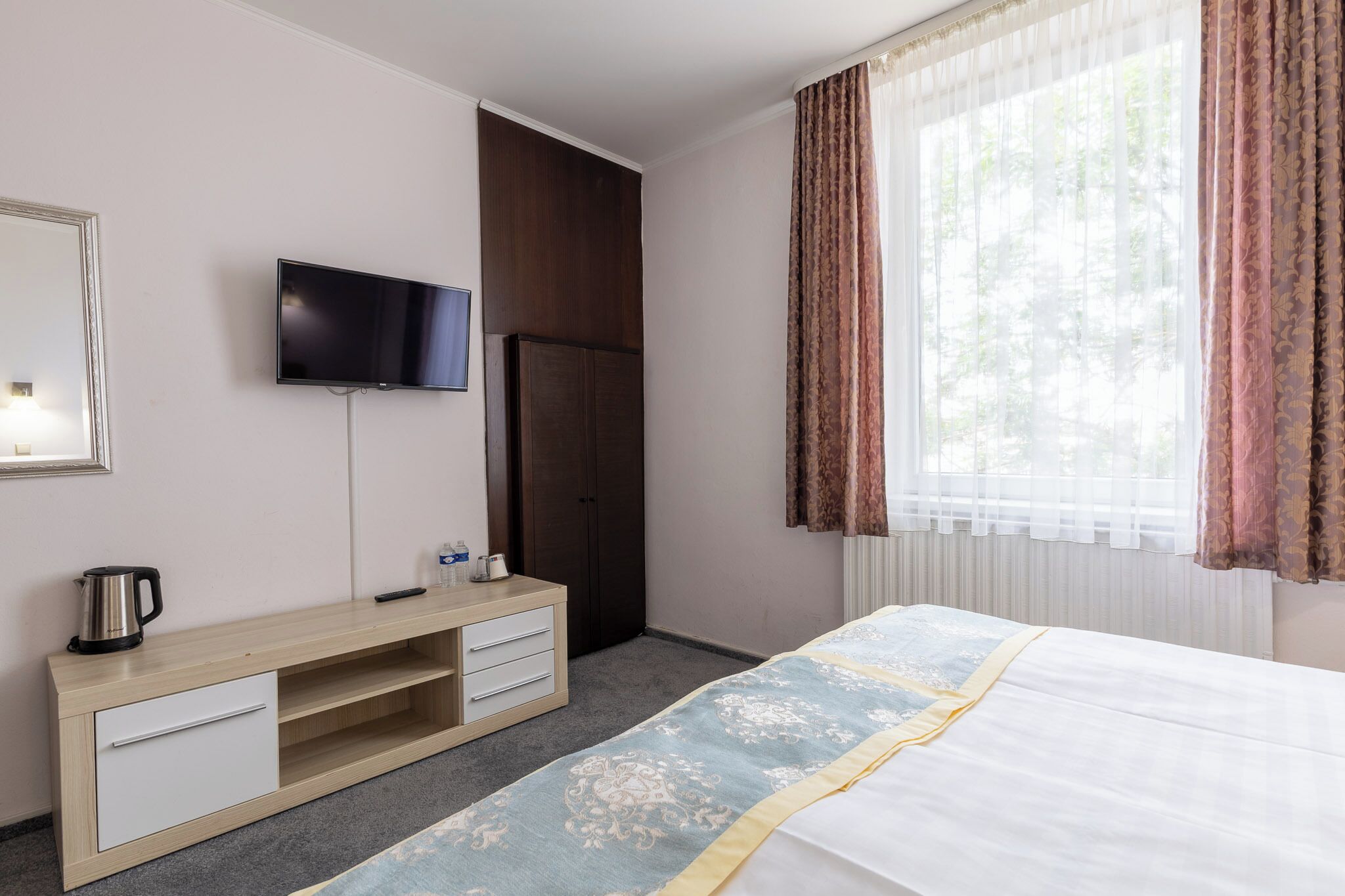 A hotel room with a bed, a TV mounted on the wall, a wooden dresser, and a window with curtains.