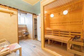 Een sauna met houten banken en een glazen deur, een klein tafeltje met een geruit kleedje, en een raam in de aangrenzende ruimte.