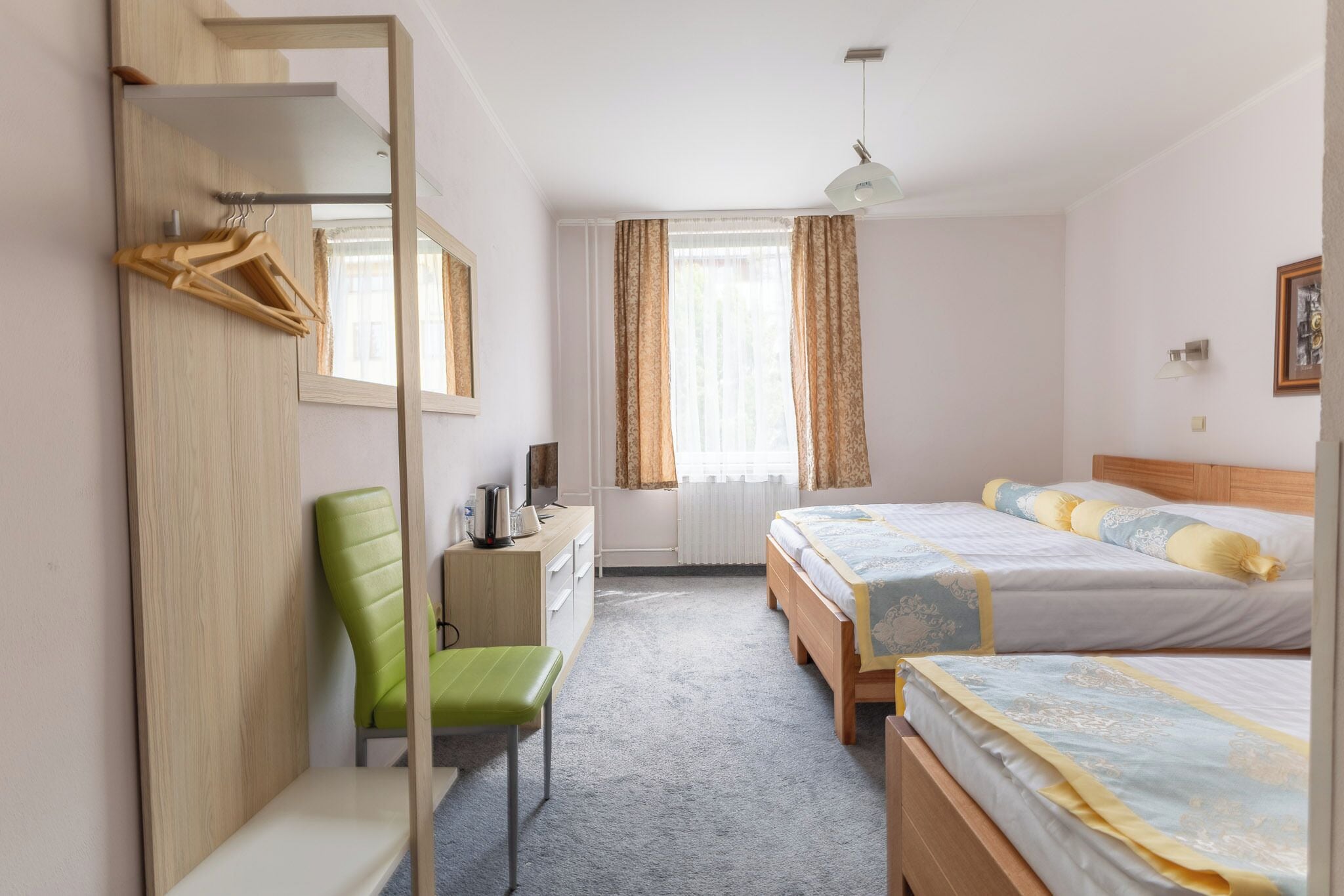 A hotel room with a wooden wardrobe, a green chair, a desk with a lamp, two beds with floral bedding, and a window with curtains.
