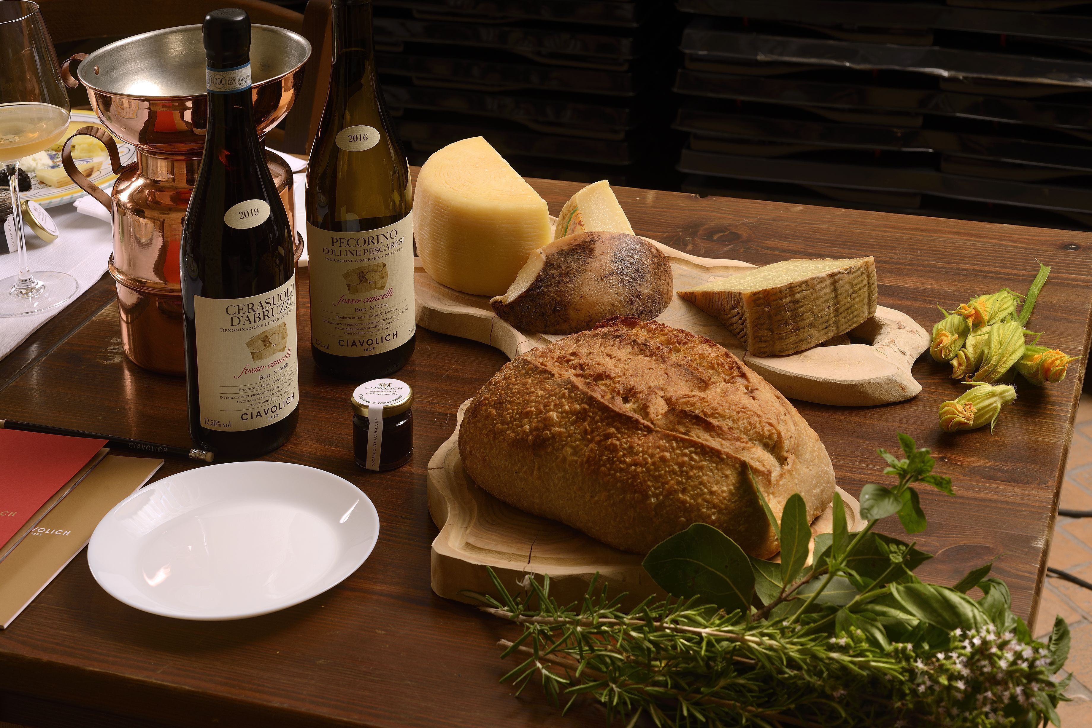 A wooden table with a bottle of wine, a cheese wheel, a loaf of bread, and some herbs.