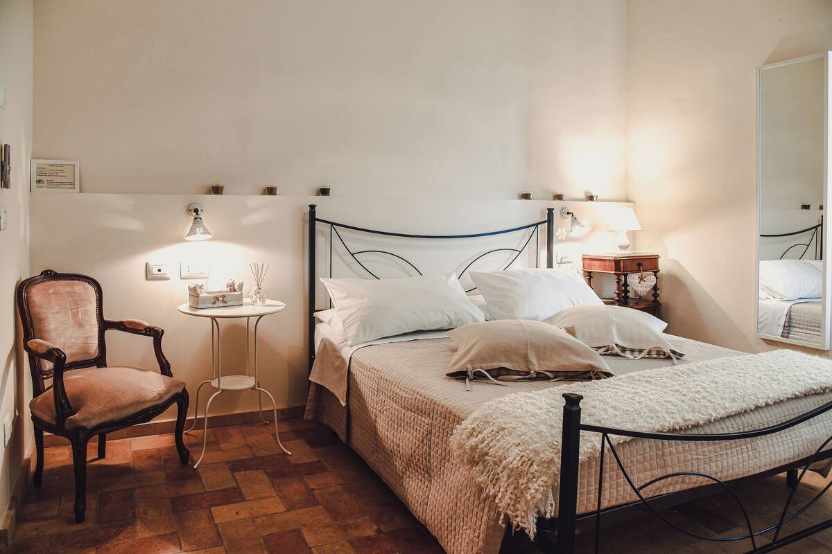 A bedroom with a bed, a chair, a bedside table, a lamp, and a mirror.