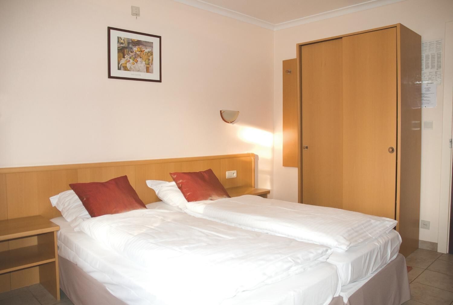Habitación de hotel con cama doble, dos almohadas, mesita de noche, lámpara fijada a la pared y armario de madera.