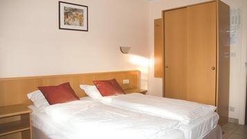 Habitación de hotel con cama doble, dos almohadas, mesita de noche, lámpara fijada a la pared y armario de madera.