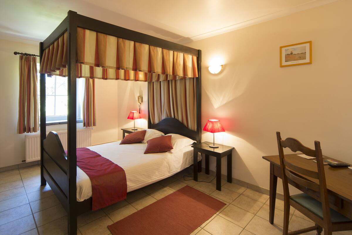 A four-poster bed with a red bedspread, two bedside tables with lamps, a wooden chair, and a framed picture on the wall.