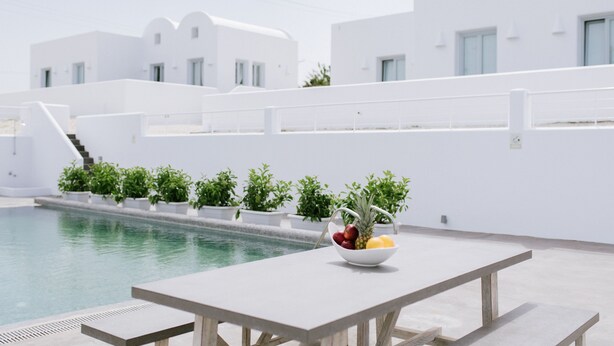 A poolside area with a table, a bowl of fruit, and white buildings in the background. - Sea and Sand Luxury Villas (Santorini)