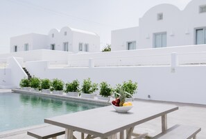 Un espace aménagé au bord de la piscine, avec une table, un saladier rempli de fruits et, en arrière-plan, des bâtiments blancs.