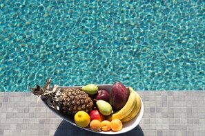A fruit bowl with pineapple, pear, apple, banana, and peach on a tiled surface by a pool. - Sea and Sand Luxury Villas (Santorini)
