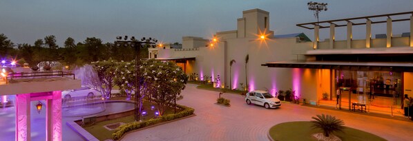A hotel entrance with a fountain, a white car parked in front, and a well-lit pathway leading to the entrance.