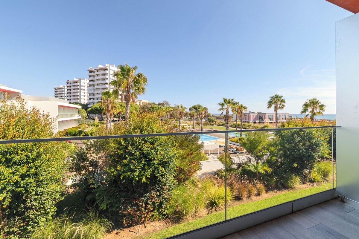 Un balcón con vistas a una piscina, abundante vegetación y un edificio distante.