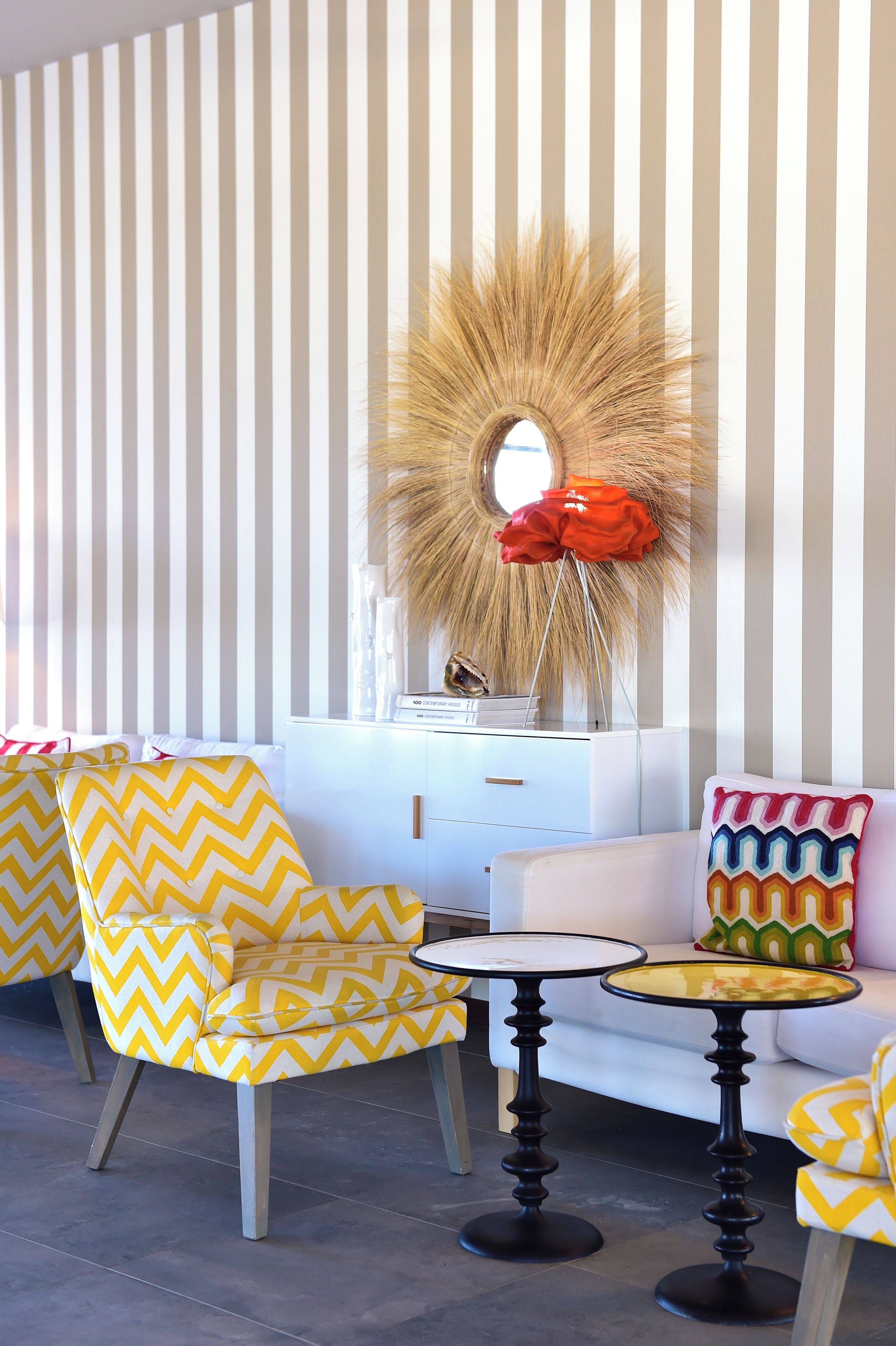 A room with striped wallpaper, a round mirror with a sunburst design, a white sofa, and two yellow chairs with zigzag patterns.