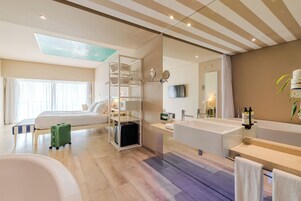 Una habitación de hotel moderna con una cama grande, un baño independiente con bañera y un espejo amplio, un escritorio pequeño y una maleta verde en el suelo.