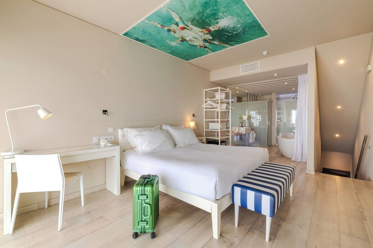 A modern hotel room with a bed, desk, and a green suitcase.