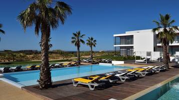 Un área de piscina con sillones de descanso y palmeras, un edificio moderno con balcones, y un cielo azul despejado.