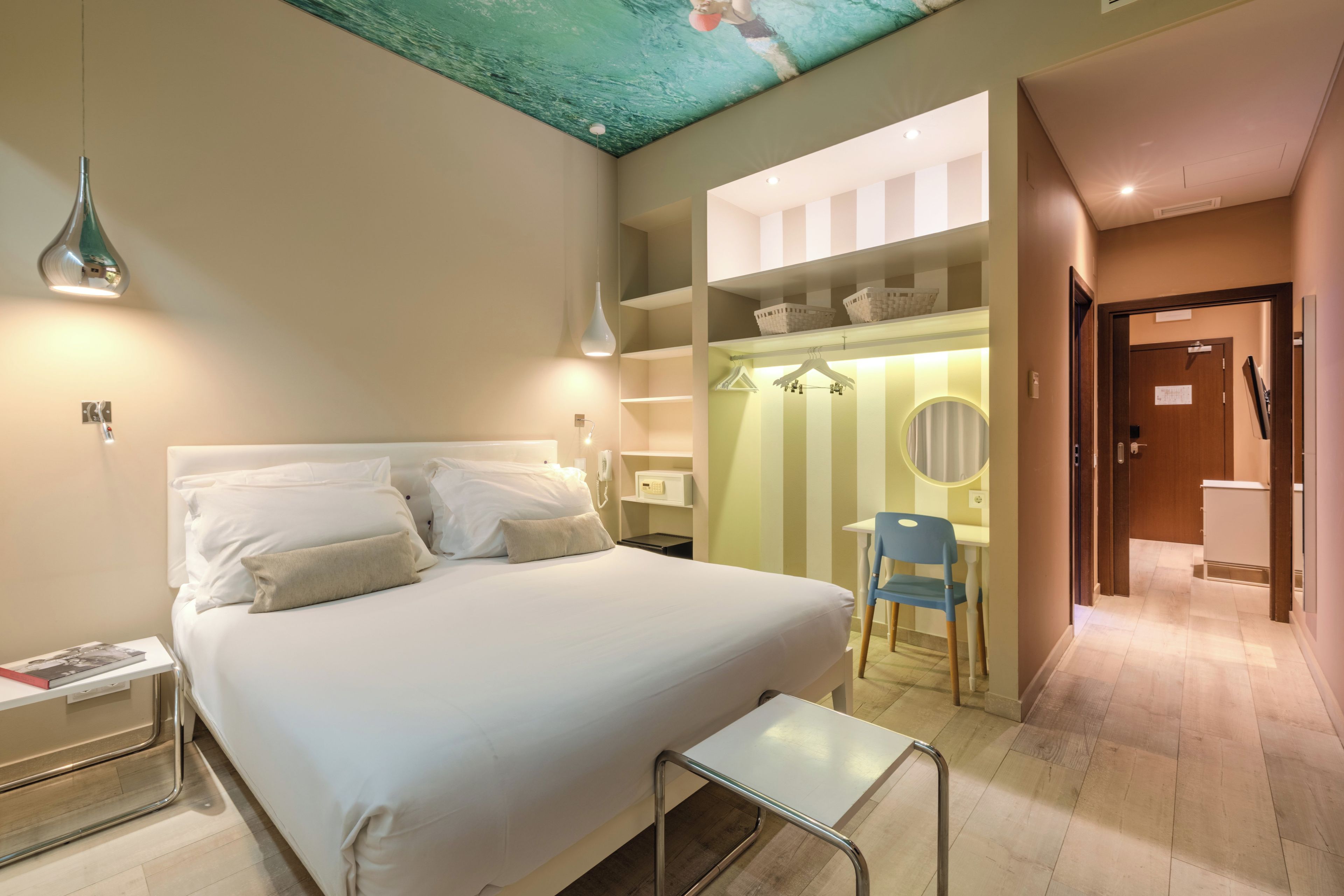 Habitación de hotel con cama, mesita de noche, lámpara, armario y un pequeño escritorio con silla.