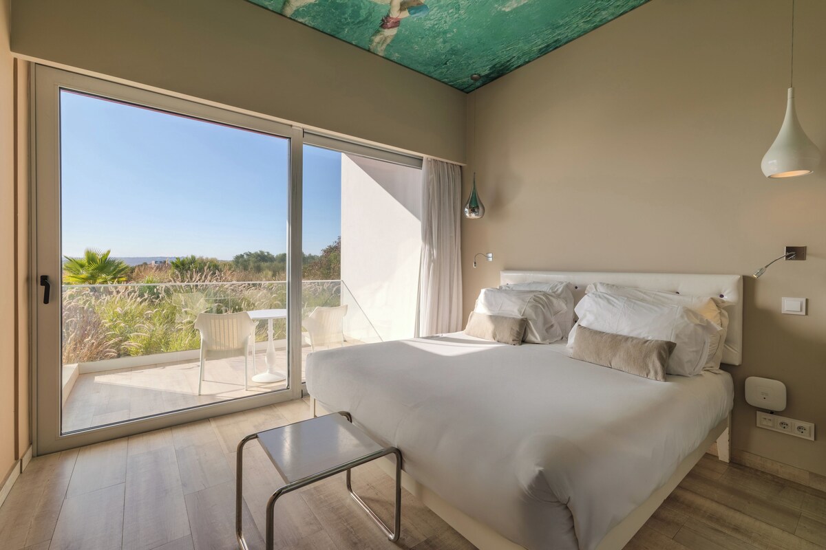 A bedroom with a large bed, a sliding glass door to a balcony, a small table, and a pendant light.