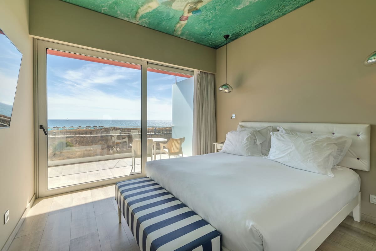 A bedroom with a large bed, a bench, a view of the sea, and a balcony with chairs.