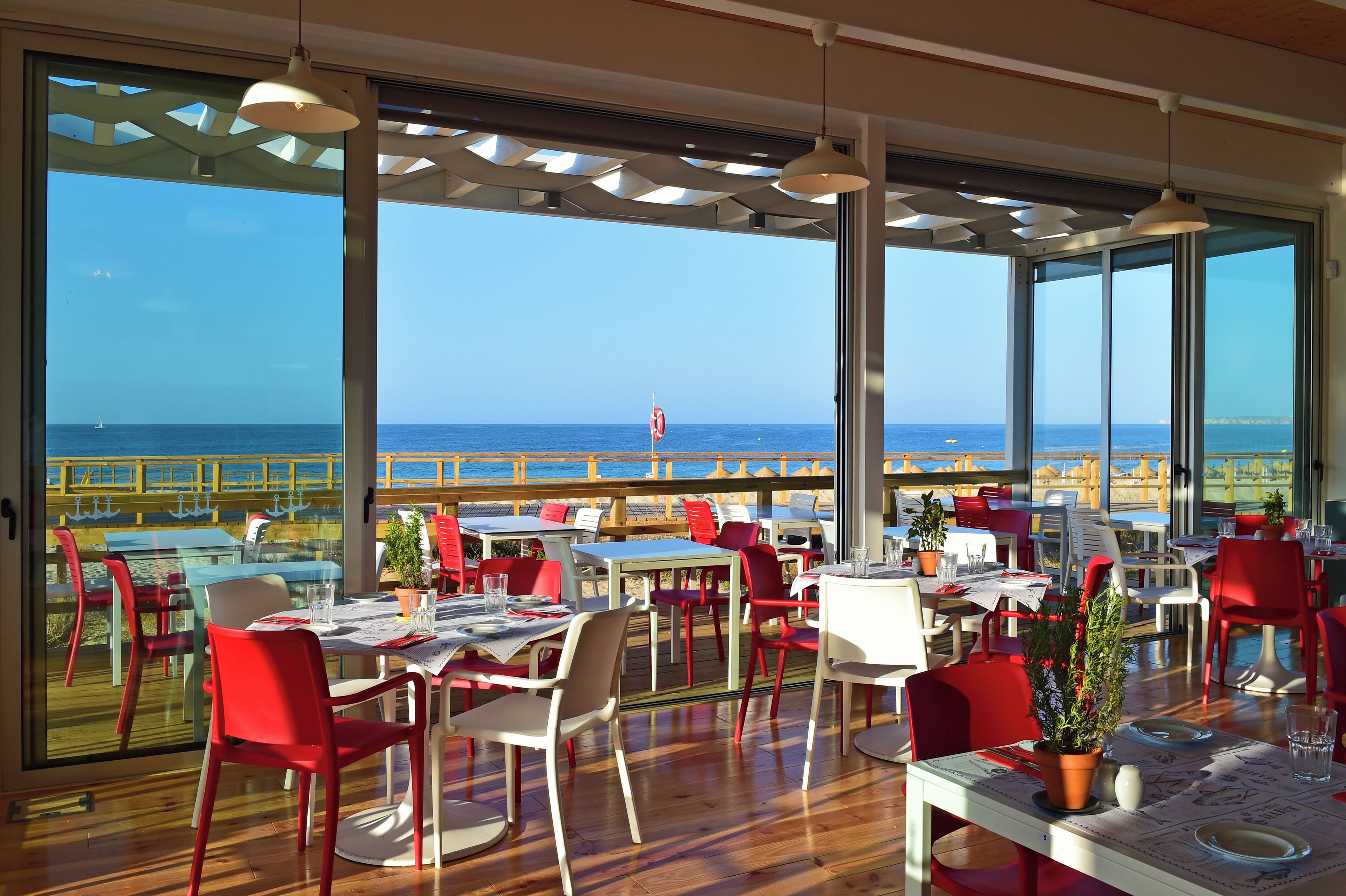 Un área de comedor al aire libre con mesas y sillas, ventanas amplias y vista al mar.