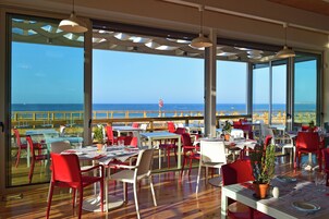 Un área de comedor al aire libre con mesas y sillas, ventanas amplias y vista al mar.