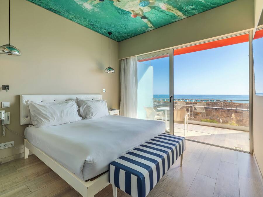 Un dormitorio con cama, un banco, un techo de diseño único y vista al océano a través de una puerta corrediza.