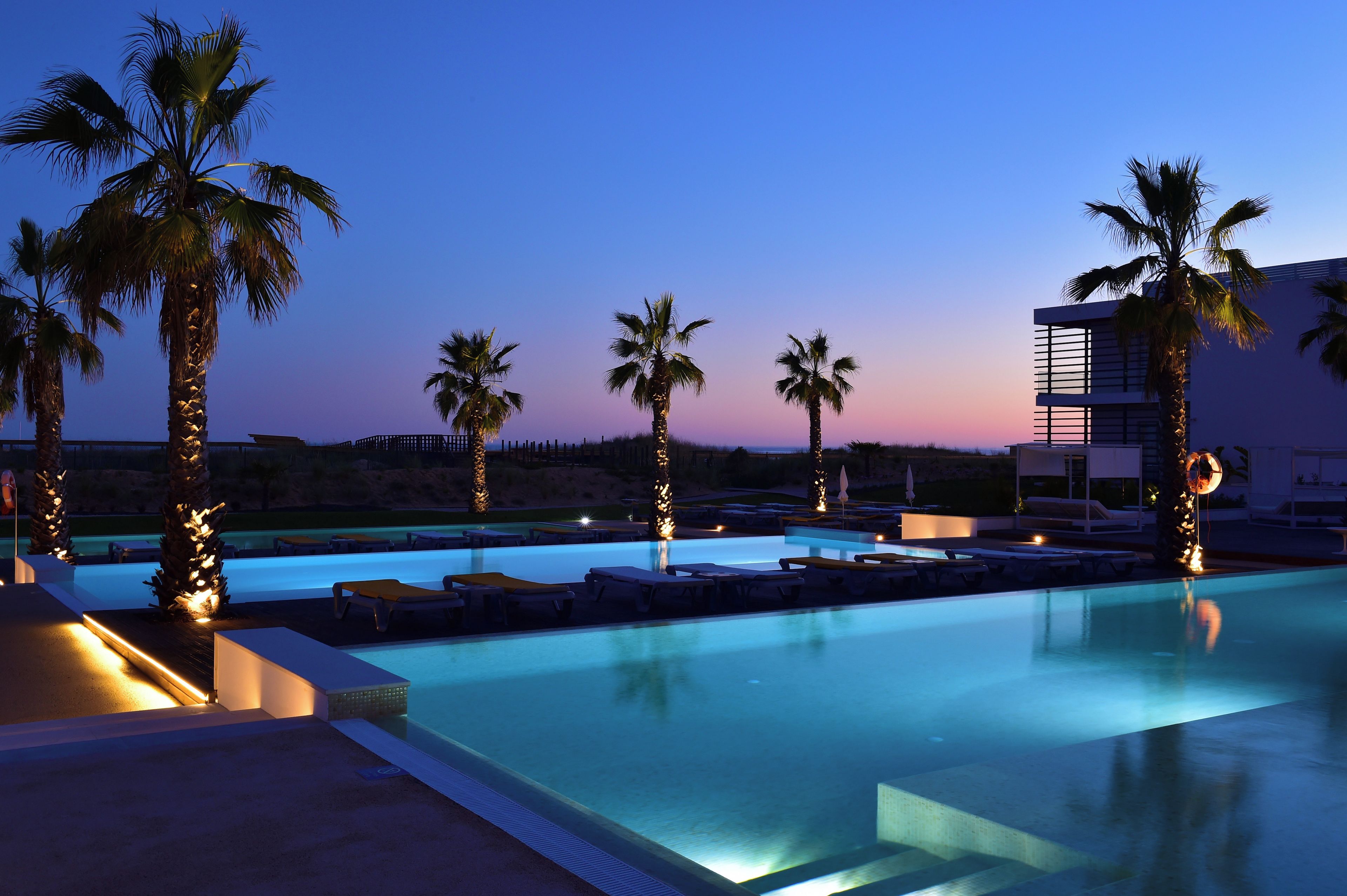 Una piscina con sillones de descanso y palmeras, rodeada por un edificio moderno.