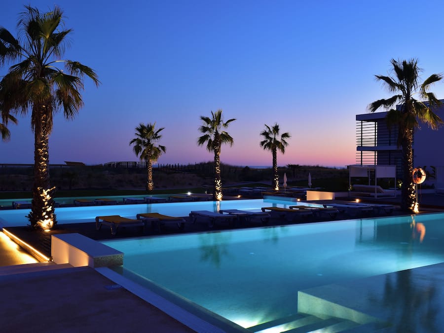 Una piscina con sillones de descanso y palmeras, rodeada por un edificio moderno.