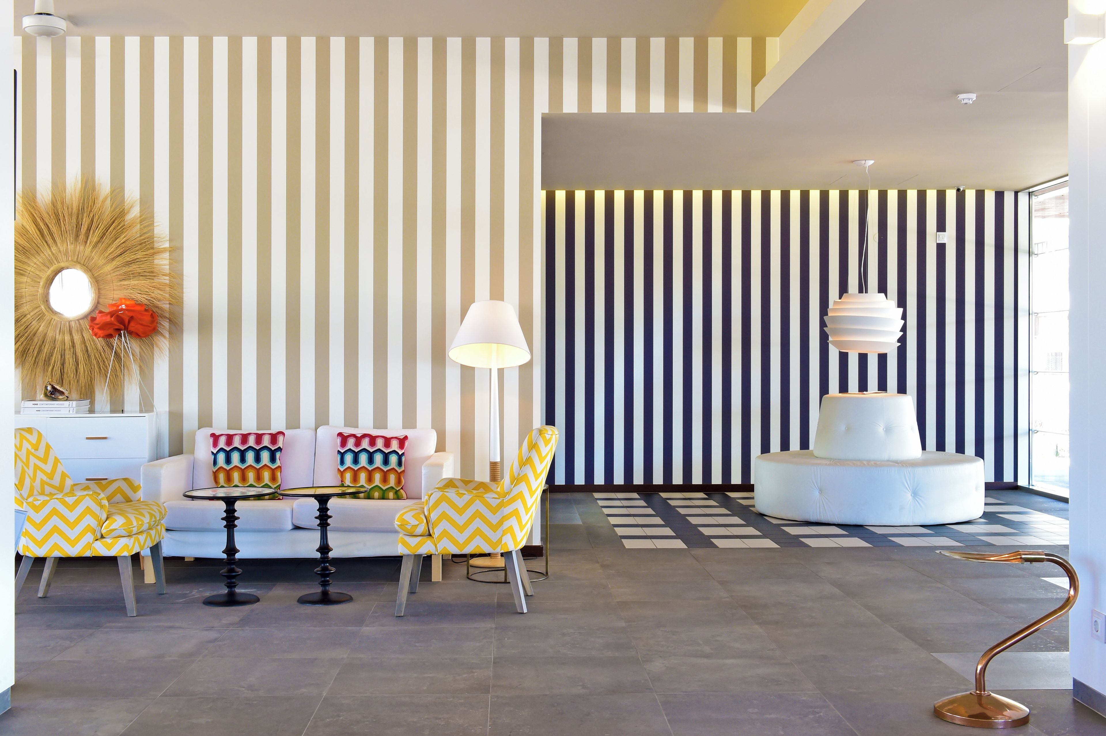 A modern living room with striped wallpaper, a round mirror, a white sofa with colorful pillows, a yellow armchair, a white table, and a unique floor lamp.