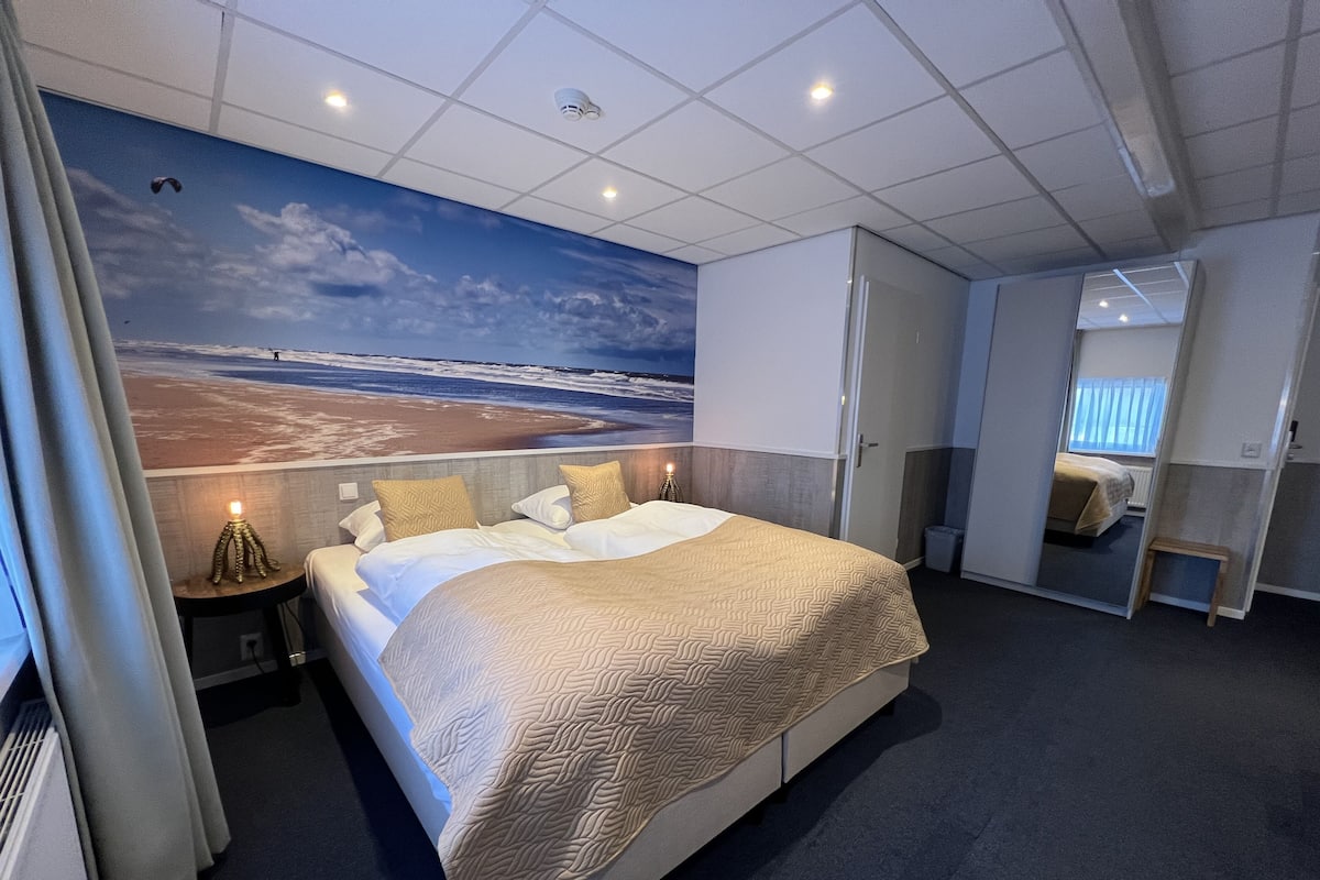 A hotel room with a large bed, a nightstand with a lamp, a wall mural of a beach, and a view of another room.