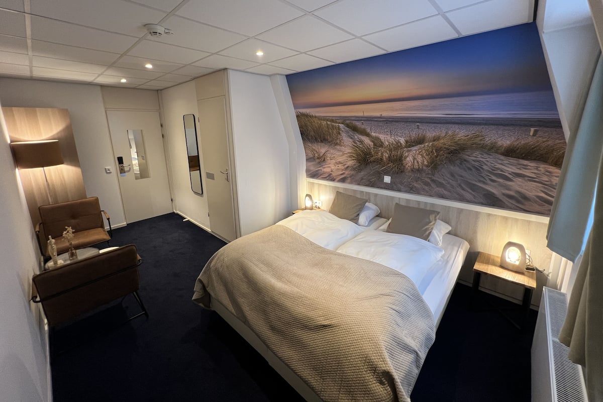 A hotel room with a bed, a desk, a chair, a mirror, and a view of the beach.