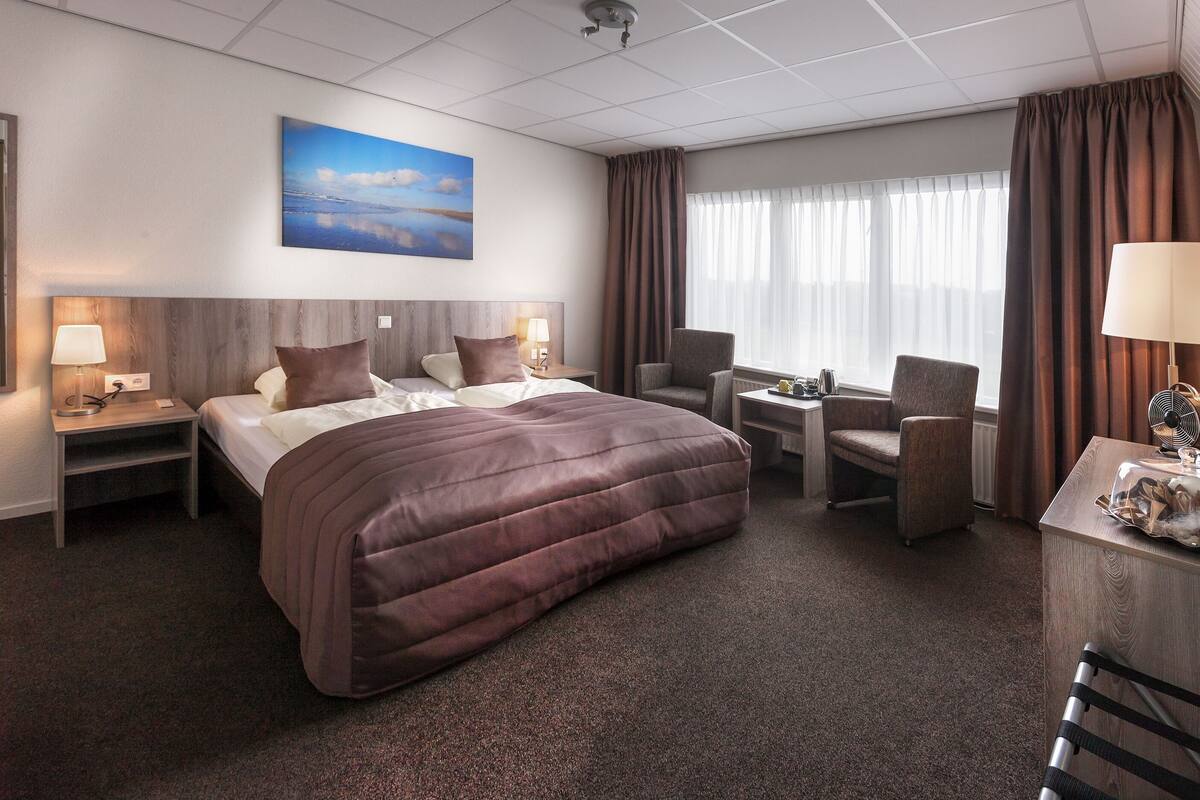 A hotel room with a large bed, two bedside tables with lamps, a desk with a chair, and a window with curtains.