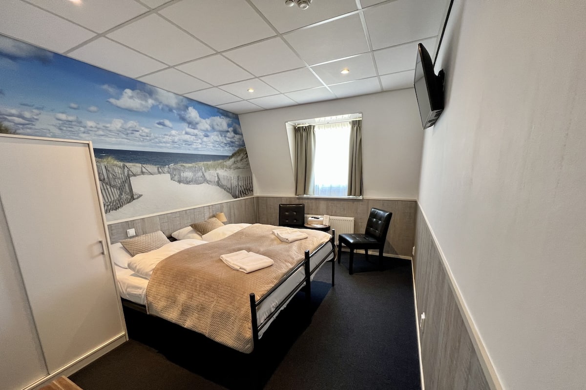 A hotel room with a bed, a desk, a chair, a TV, and a mural of a beach scene.