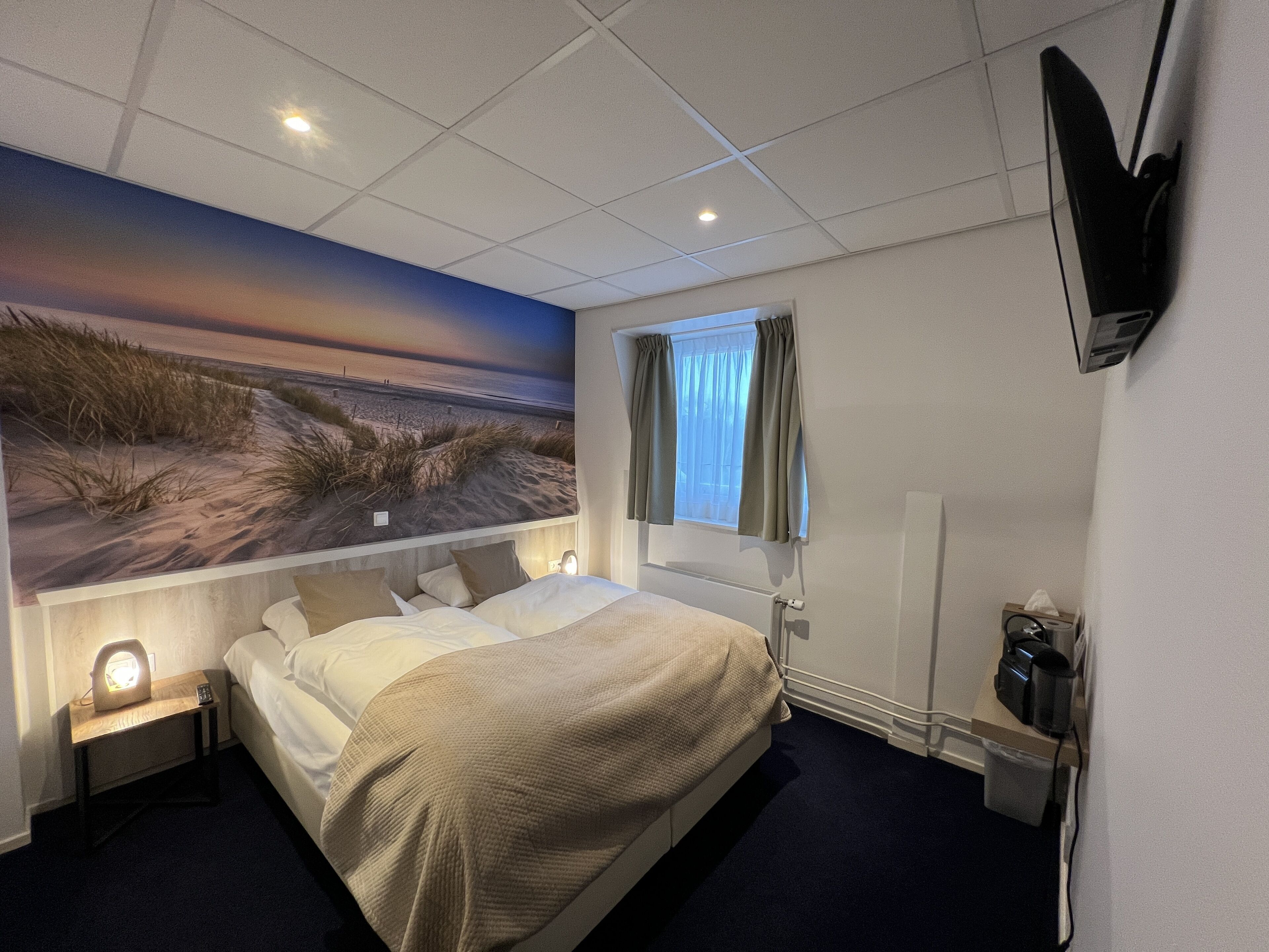 Una habitación de hotel con cama, mesita de noche, televisor montado en la pared y ventana con cortinas.