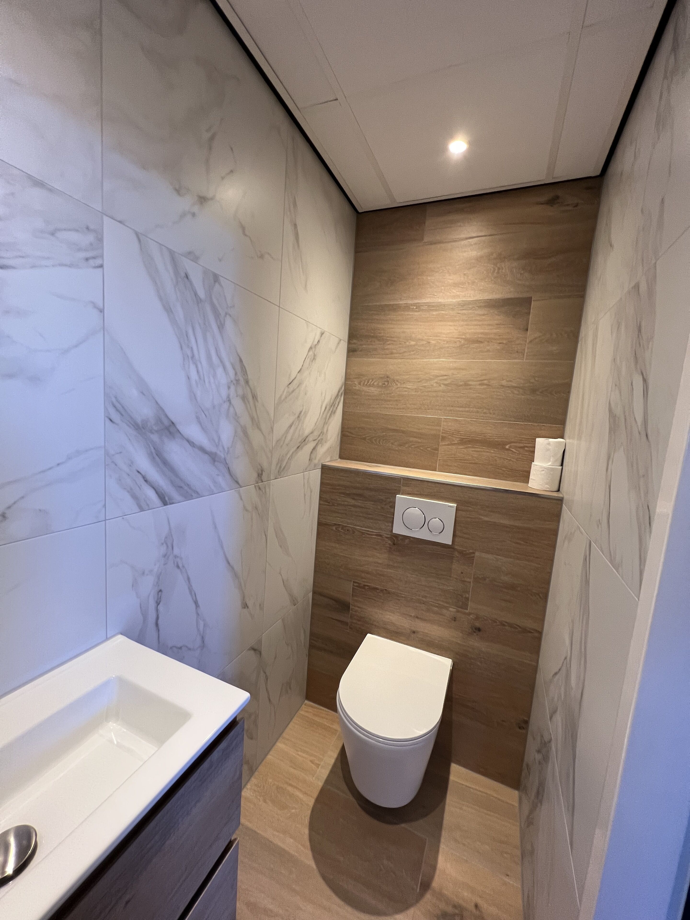 A modern bathroom with marble walls, a wooden floor, a white sink, and a toilet.