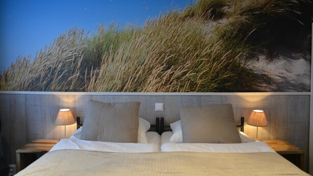 Een bed met twee kussens, een nachtkastje met een lamp en een muur met een grasachtige textuur.