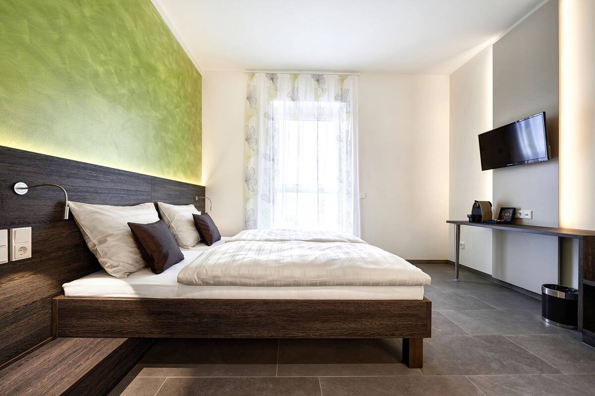 A modern bedroom with a wooden headboard, a flat-screen TV mounted on the wall, a bedside table with a lamp, and a large window with sheer curtains.