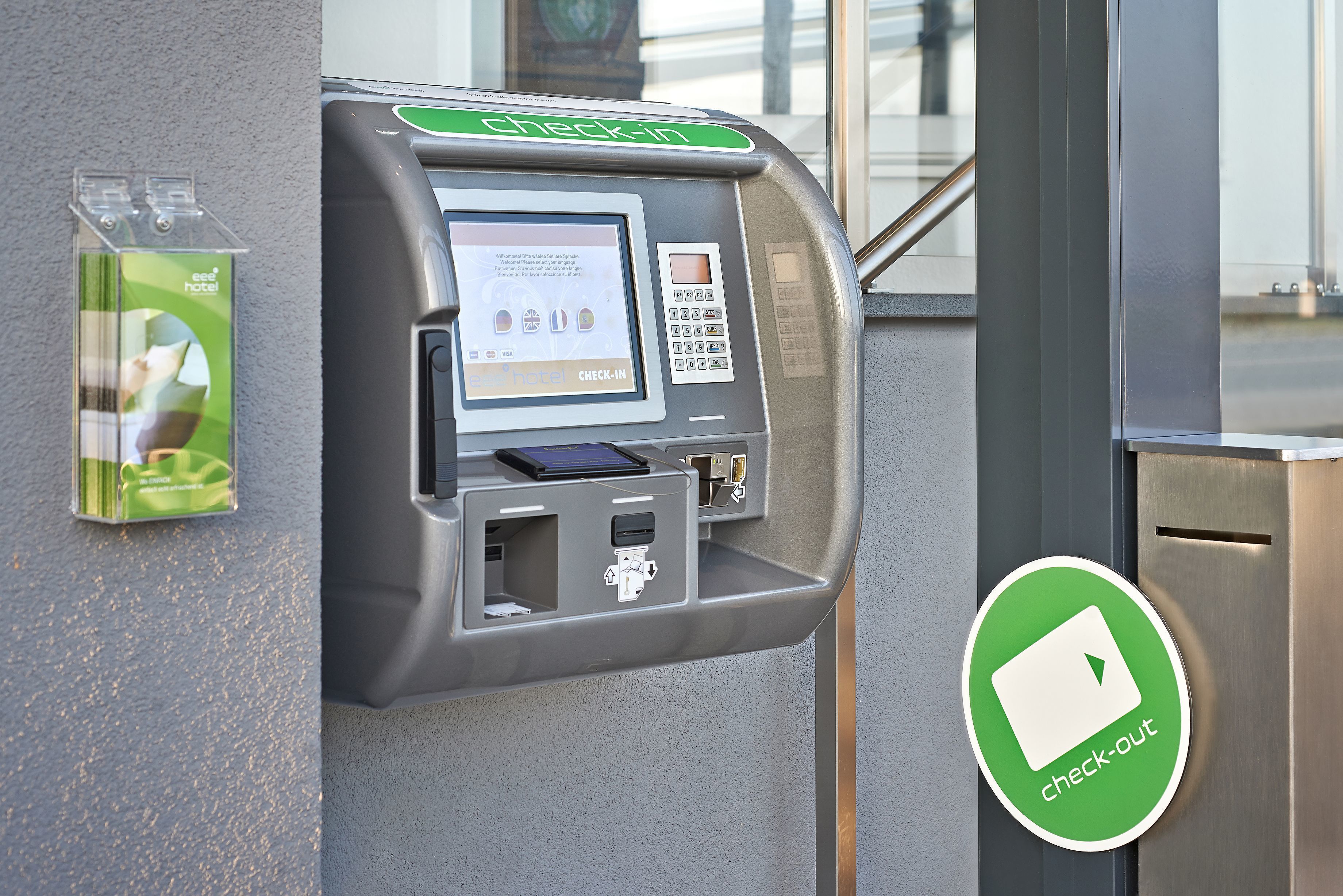 A check-in kiosk with a digital screen, a card reader, and a green sign indicating check-out.