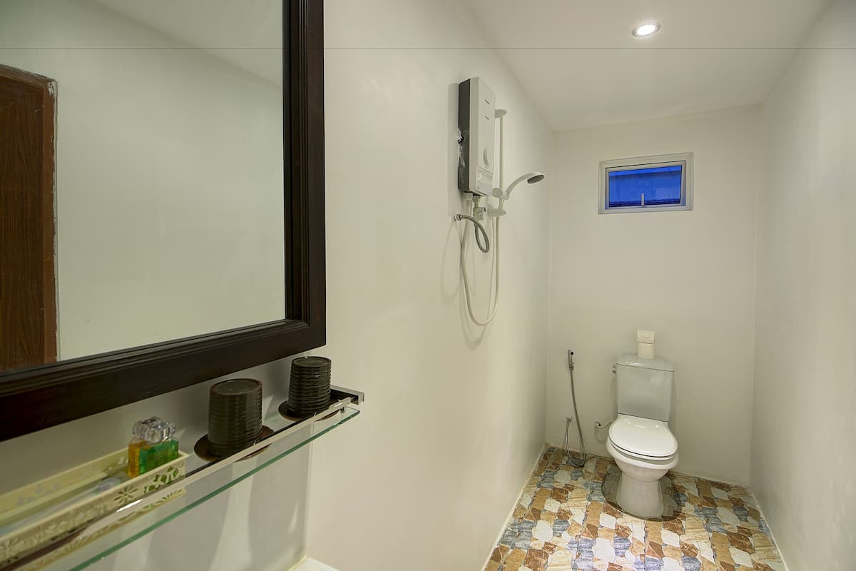 A small bathroom with a toilet, a shower, a glass shelf with toiletries, and a window.