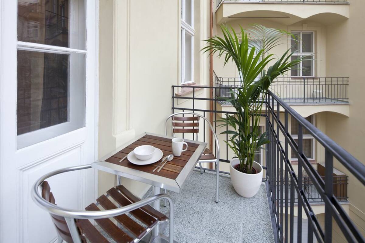 Un balcón con una mesa para dos personas, una planta en maceta y vista al interior del edificio.