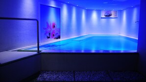 A blue-lit indoor swimming pool with a tiled floor, a bench, and a wall-mounted TV.