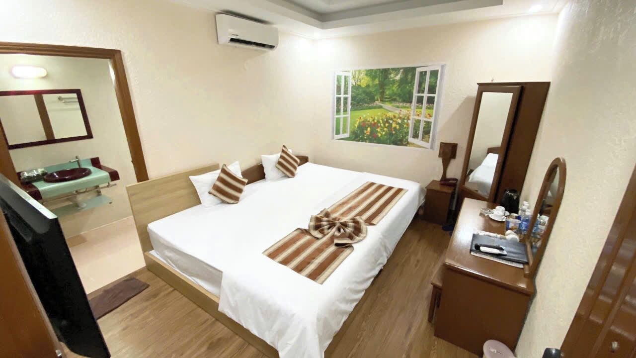 A hotel room with a bed, a bedside table, a mirror, a window with a view of greenery, and a bathroom with a sink and mirror.