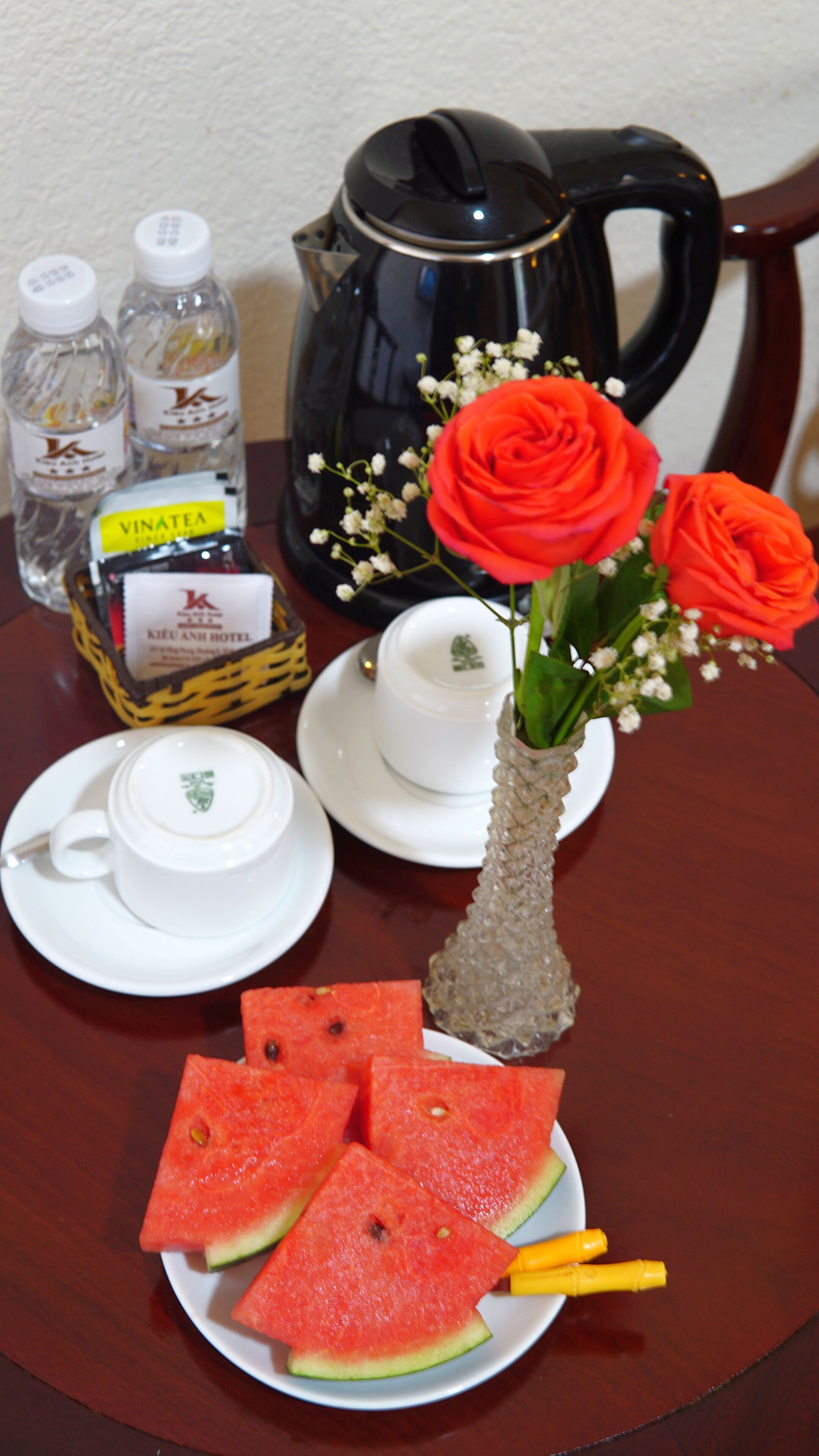Ein Tisch mit Teekanne, zwei Flaschen, einer Rosenvase, einer Tasse, einem Untertasse und Wassermelonenscheiben.
