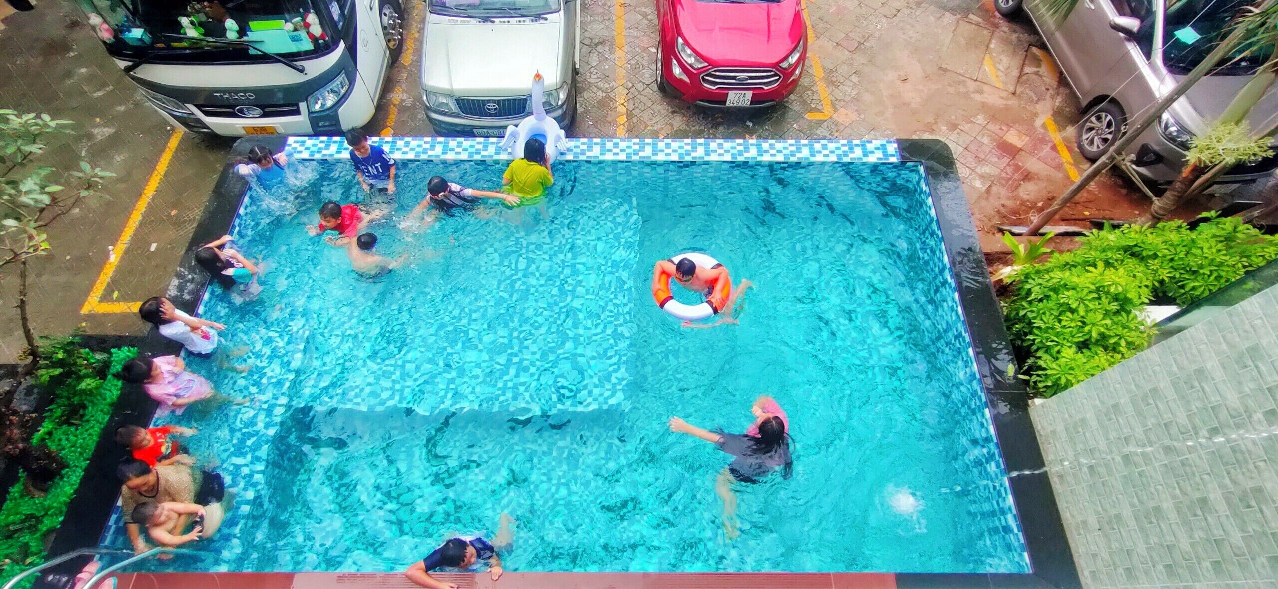 A swimming pool with people swimming and playing, surrounded by parked cars and greenery.